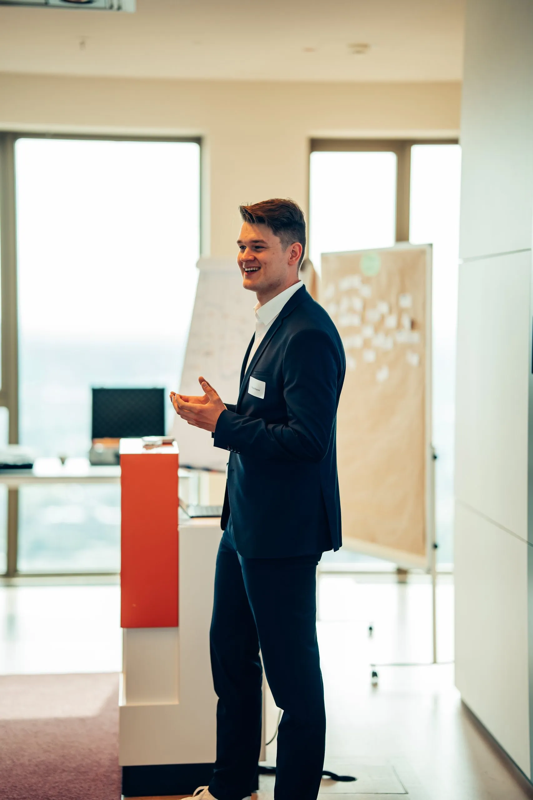 Pascal Kayser spricht lächelnd im Workshop und gestikuliert, hinter ihm Flipchart, Pinnwand mit Haftnotizen und helle Fenster
